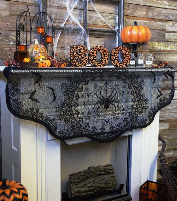 A Halloween-themed mantel features a black lace spiderweb cover with bat motifs. Decor includes "BOO" letters, candles, pumpkins, and faux cobwebs, evoking a spooky yet playful atmosphere.