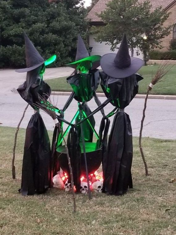 Three witch figures in black hats stand around a cauldron glowing green. Skulls and red lights are placed underneath, creating a spooky scene.