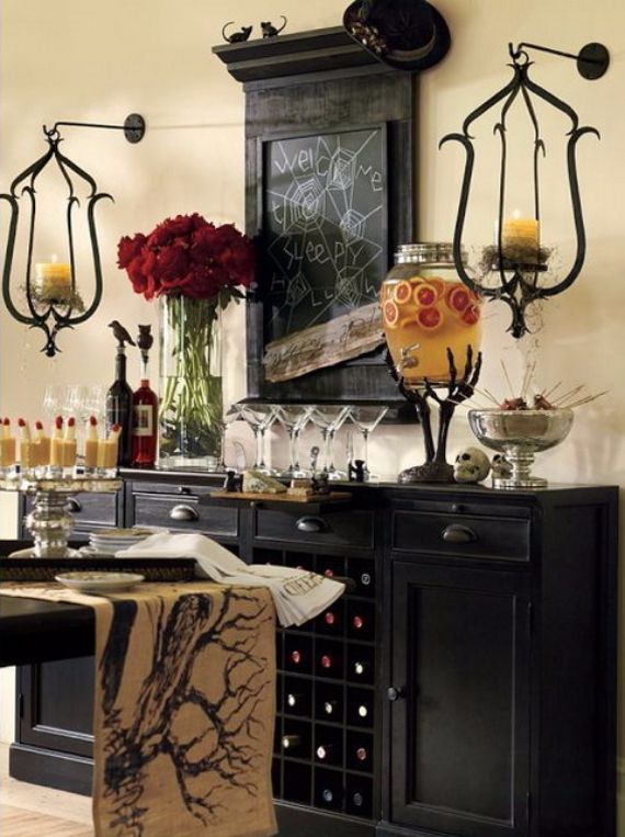 A Halloween-themed dining area features a black sideboard with red roses, wine glasses, a skull, and a large jar of fruit punch. A chalkboard with "Welcome Sleepy Hollow" in white adds an eerie touch.