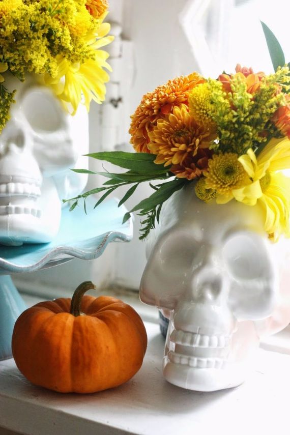 White ceramic skulls serve as vases for vibrant orange and yellow flowers, beside a small pumpkin on a windowsill, creating a festive autumn display.