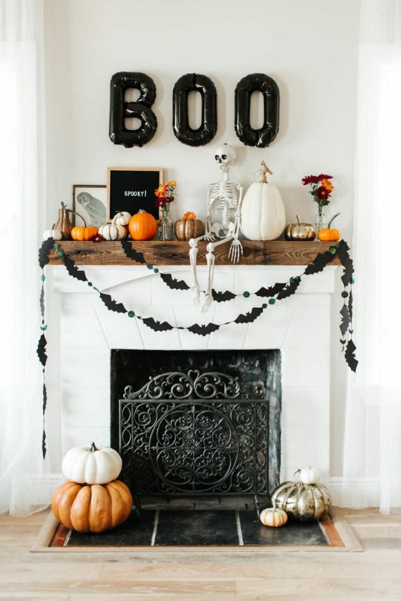 Halloween-themed fireplace with a wooden mantel adorned with pumpkins, a small skeleton, and "BOO" balloons. Bat garlands hang festively below.