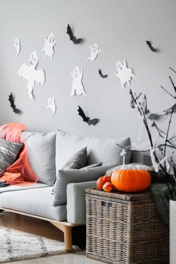 A cozy Halloween-themed living room with white ghost and black bat decorations on a gray wall. A gray sofa with orange and gray cushions sits below, next to a wicker basket adorned with pumpkins. Branches with cobwebs add a spooky touch.