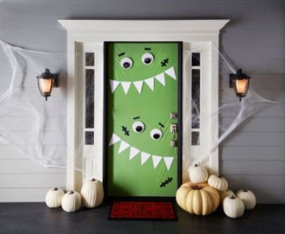 The front door is decorated like a green monster with large eyes and sharp white teeth for Halloween. White pumpkins and cobwebs add spooky charm.