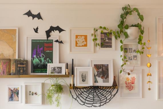 A cozy wall display features framed art, a shelf with books, and a hanging plant. Black paper bats and a web add a playful, Halloween touch.