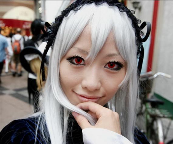 A person cosplays with long silver hair, striking red eyes, and an intricate black headpiece, smiling softly. Background shows a blurred street scene.