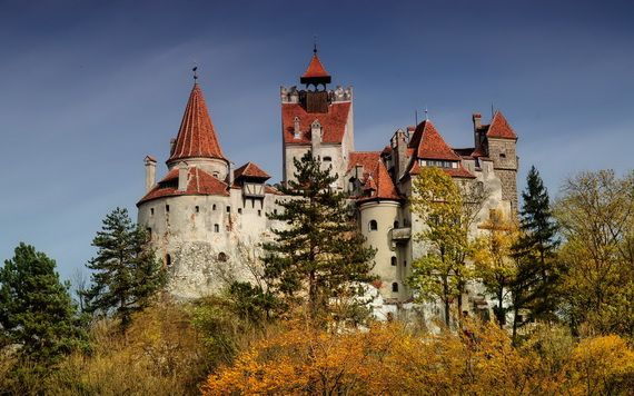 A majestic medieval castle with red-tiled roofs and turrets stands against a clear blue sky, surrounded by lush green and autumnal trees.
