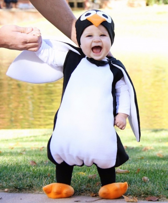 A joyful child in a penguin costume, with orange feet and a beak, stands on grass. An adult's hands support them. The background shows a pond.