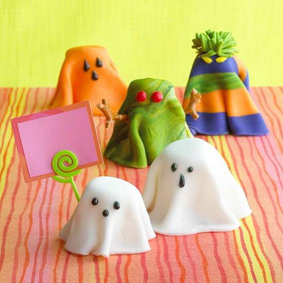 Playful fondant ghosts and monsters on a colorful striped tablecloth. Two white ghosts in front, and bright orange, green, and striped creatures behind.