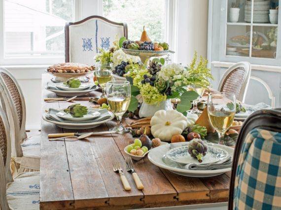 Rustic dining table set with floral centerpiece, figs, green and white pumpkins, wine glasses, and plates, creating an inviting, autumn ambiance.