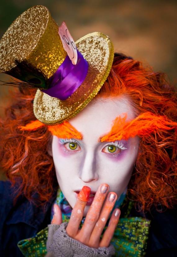 A person dressed as a whimsical character, with white face paint, bright orange hair and eyebrows, wearing a glittery gold hat with a purple band, conveys a playful and eccentric vibe.
