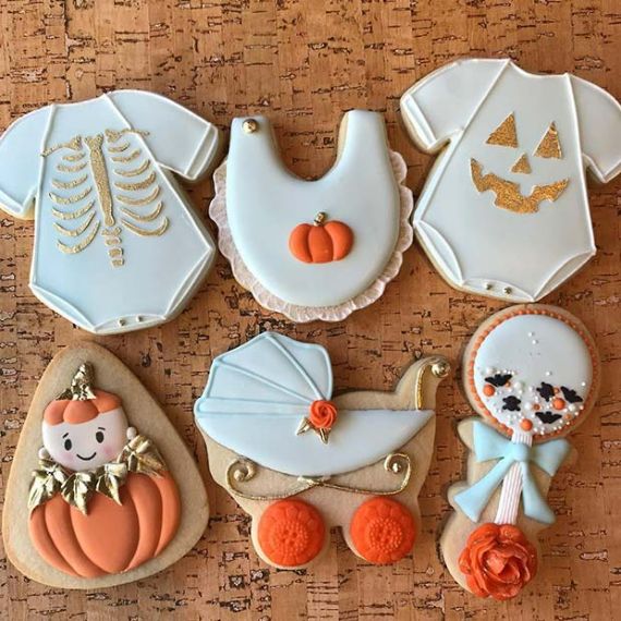 Halloween-themed cookies on a cork background. They include a ribcage, jack-o'-lantern, pumpkin bib, baby carriage, pumpkin baby, and skeleton girl. Festive and playful.