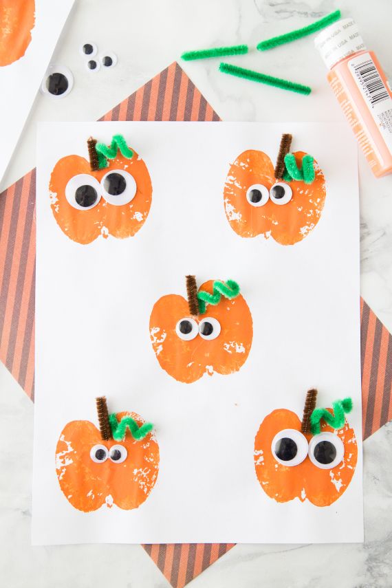 Five playful pumpkin stamps with googly eyes and green pipe cleaner stems are arranged on white paper, evoking a fun, crafty Halloween vibe.