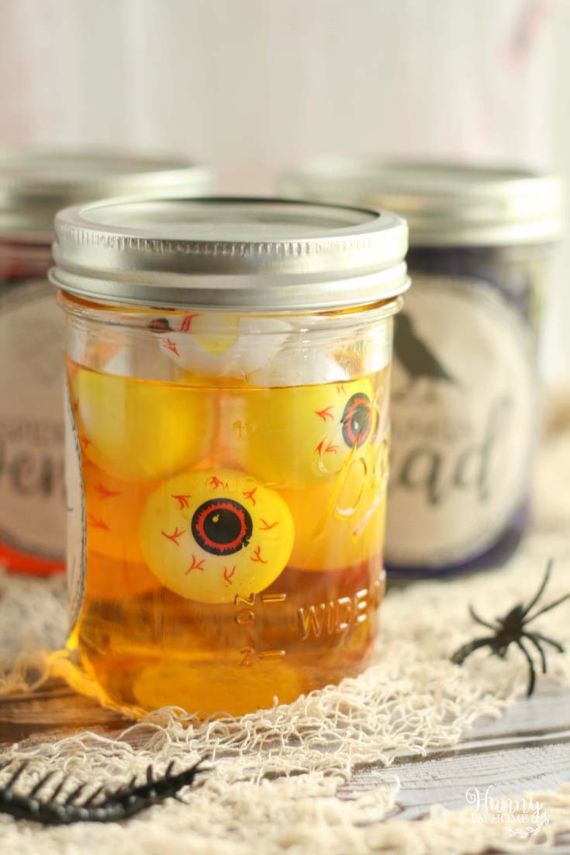 A jar filled with yellow eyeball-shaped candies in liquid, with Halloween decor. The background shows blurred jars and fake spiders on a spooky cloth.